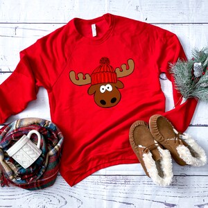 May include: Red long-sleeved sweatshirt with a moose wearing a red and white knit hat with a white pom-pom. The moose has brown antlers and a brown face.