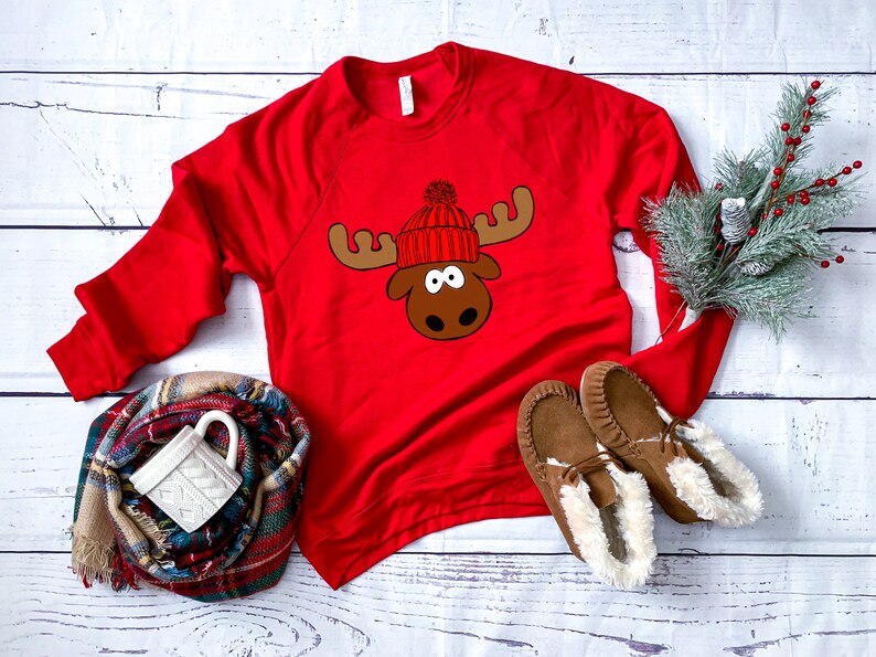 May include: Red long-sleeved sweatshirt with a moose wearing a red and white knit hat with a white pom-pom. The moose has brown antlers and a brown face.