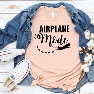 May include: A peach-colored t-shirt with the text "Airplane Mode" in black lettering. A small airplane silhouette is flying above the text with a dashed line behind it.