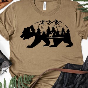 May include: A brown t-shirt with a black silhouette of a bear with a mountain range, trees, and a deer inside the bear's outline.