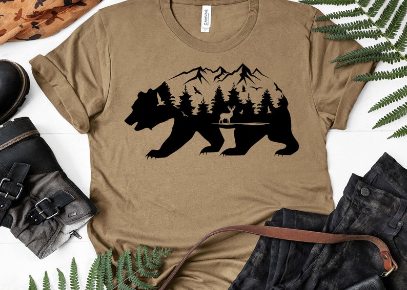May include: A brown t-shirt with a black silhouette of a bear with a mountain range, trees, and a deer inside the bear's outline.
