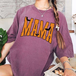 May include: A mauve-colored t-shirt with the word "MAMA" in orange block letters, accented with black bat silhouettes. The shirt has a relaxed fit and is suitable for Halloween or everyday wear.