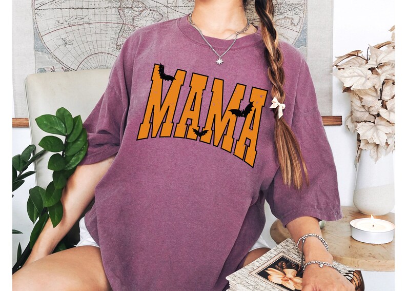 May include: A mauve-colored t-shirt with the word "MAMA" in orange block letters, accented with black bat silhouettes. The shirt has a relaxed fit and is suitable for Halloween or everyday wear.