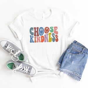 May include: White t-shirt with the text "CHOOSE KINDNESS" in colorful retro font with smiley faces. The shirt is laid flat on a white surface with a pair of gray sneakers and blue denim shorts.