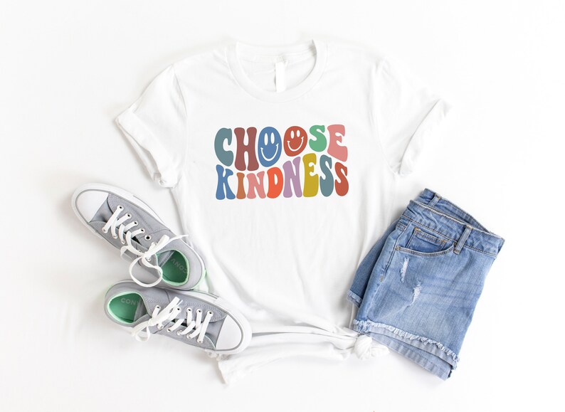 May include: White t-shirt with the text "CHOOSE KINDNESS" in colorful retro font with smiley faces. The shirt is laid flat on a white surface with a pair of gray sneakers and blue denim shorts.