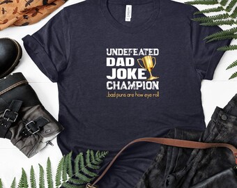Op de afbeelding: Een donkerblauw T-shirt met witte tekst die "Undefeated Dad Joke Champion" luidt en een gouden trofee-afbeelding. De tekst onder de trofee luidt "Bad puns are how eye roll."