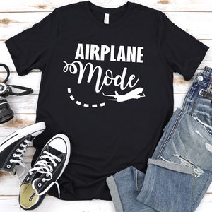 May include: Black t-shirt with white text that says "Airplane Mode" and a white airplane graphic. The shirt is laid out on a white wooden surface with a pair of black sneakers, a pair of blue jeans, and a camera.