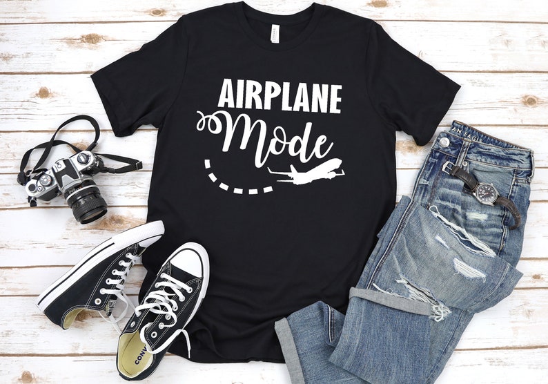 May include: Black t-shirt with white text that says "Airplane Mode" and a white airplane graphic. The shirt is laid out on a white wooden surface with a pair of black sneakers, a pair of blue jeans, and a camera.