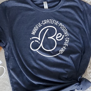 May include: A navy blue t-shirt with a white circle design that says "Be" in a stylized font. The circle is surrounded by the words "Mindful, Grateful, Positive, True, Kind".