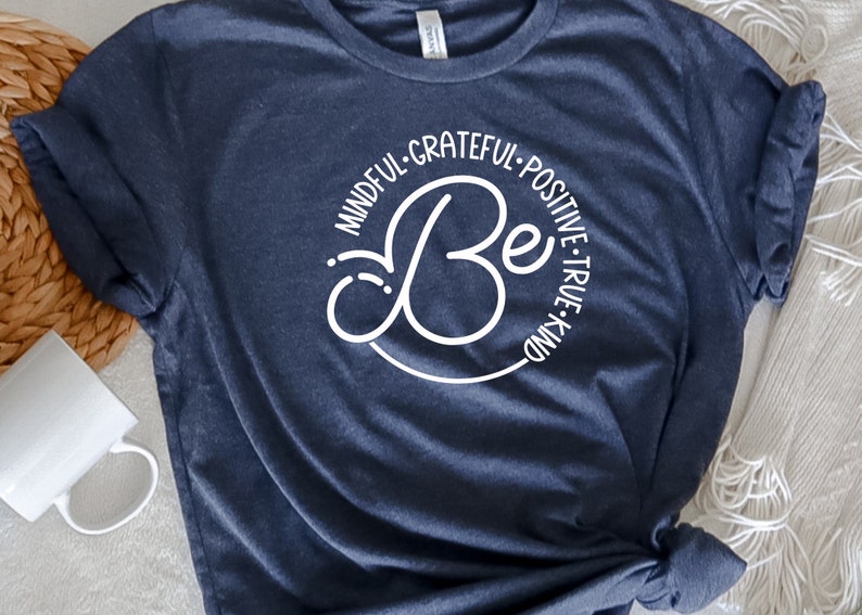 May include: A navy blue t-shirt with a white circle design that says "Be" in a stylized font. The circle is surrounded by the words "Mindful, Grateful, Positive, True, Kind".