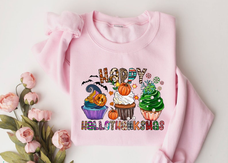 K&ouml;nnte beinhalten: Ein hellrosa Sweatshirt mit den Worten "HAPPY HALLOTHANKSMAS" in bunten Buchstaben. Das Design zeigt drei Cupcakes, die mit K&uuml;rbissen, Flederm&auml;usen und Schneeflocken verziert sind. Die Cupcakes haben unterschiedlich farbige Glasuren und Dekorationen.