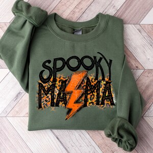 May include: Olive green sweatshirt with the words "Spooky Mama" in black, with an orange lightning bolt and leopard print accents. The design includes a spiderweb. A casual, comfortable top for Halloween.