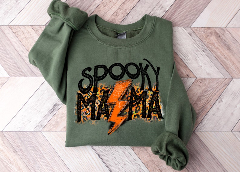 May include: Olive green sweatshirt with the words "Spooky Mama" in black, with an orange lightning bolt and leopard print accents. The design includes a spiderweb. A casual, comfortable top for Halloween.
