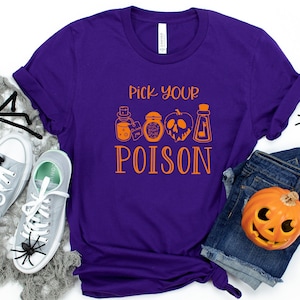 May include: Purple t-shirt with the text "Pick Your Poison" in orange, featuring potion bottle illustrations. The shirt is styled with gray sneakers, a denim skirt, and Halloween decorations including spiders and a carved pumpkin.