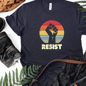 May include: A navy blue t-shirt with a vintage-style graphic of a raised fist in front of a sunset. The text "RESIST" is printed below the graphic.
