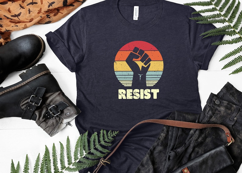 May include: A navy blue t-shirt with a vintage-style graphic of a raised fist in front of a sunset. The text "RESIST" is printed below the graphic.