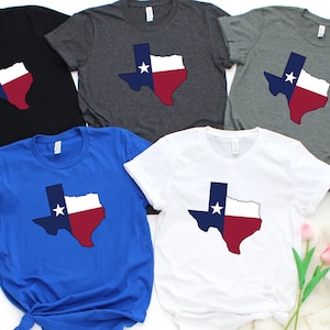 Op de afbeelding: Vier T-shirts met een ontwerp van de omtrek van de staat Texas met de kleuren van de Texas-vlag: blauw, wit en rood. De shirts zijn in zwart, grijs melange, koninklijk blauw en wit.