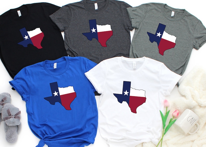 Op de afbeelding: Vier T-shirts met een ontwerp van de omtrek van de staat Texas met de kleuren van de Texas-vlag: blauw, wit en rood. De shirts zijn in zwart, grijs melange, koninklijk blauw en wit.