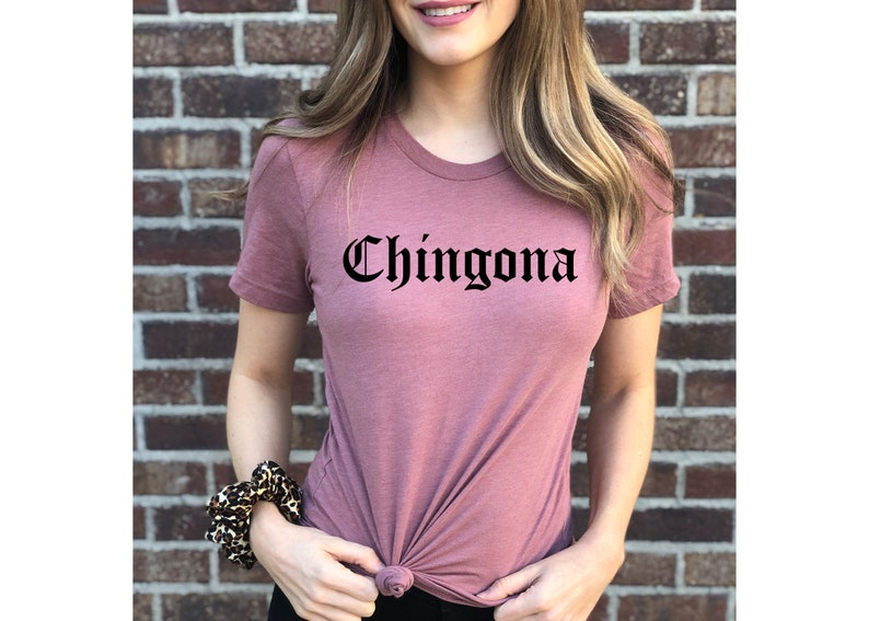 May include: A mauve t-shirt with the word "Chingona" printed in black gothic font.