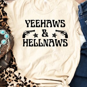May include: A beige t-shirt with the text "Yeehaws & Hellnaws" in black lettering. Two black silhouettes of pistols are on either side of the text, with a black star on each side of the ampersand.