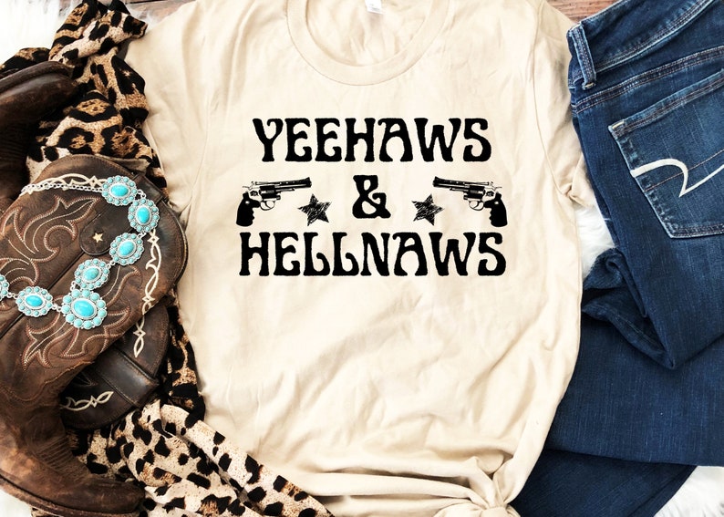 May include: A beige t-shirt with the text "Yeehaws & Hellnaws" in black lettering. Two black silhouettes of pistols are on either side of the text, with a black star on each side of the ampersand.