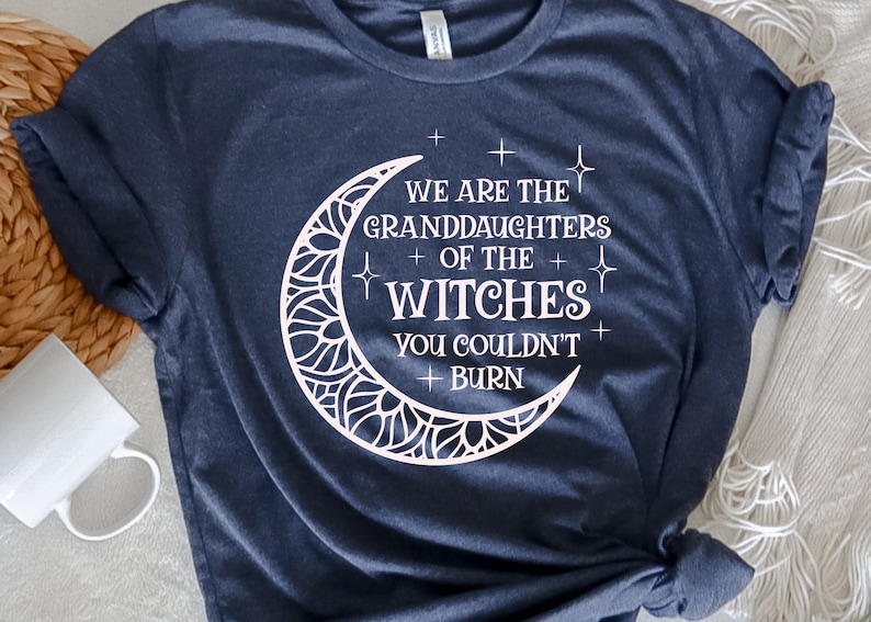 May include: A dark blue t-shirt with a white crescent moon design. The moon has a decorative pattern and the text "We are the granddaughters of the witches you couldn't burn" is written inside the moon.