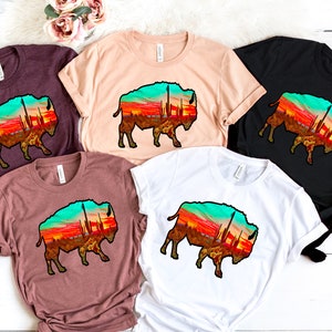 May include: Four t-shirts with a graphic of a silhouette of a bison with a sunset and cacti inside the silhouette. The t-shirts are in maroon, beige, black, and white.