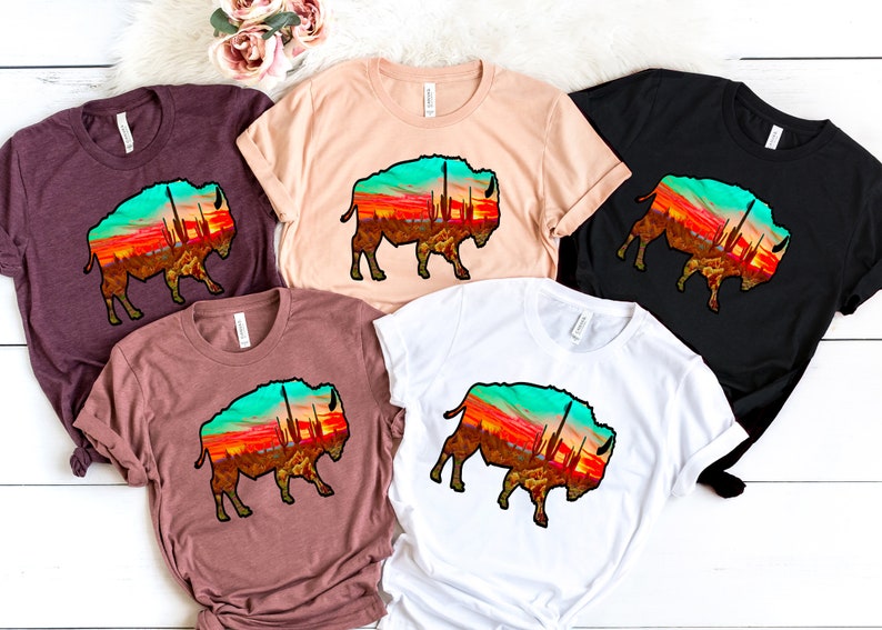 May include: Four t-shirts with a graphic of a silhouette of a bison with a sunset and cacti inside the silhouette. The t-shirts are in maroon, beige, black, and white.