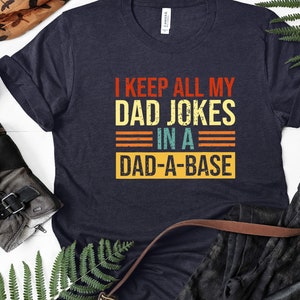 May include: A dark blue t-shirt with a vintage-style graphic that reads "I keep all my dad jokes in a dad-a-base".