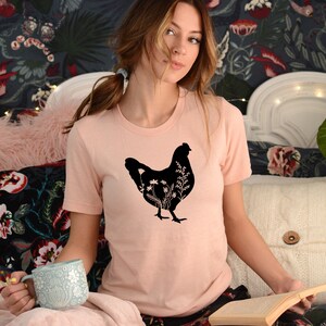 Pode incluir: Uma camiseta rosa com uma silhueta preta de uma galinha com flores dentro. A galinha est&aacute; virada para a direita e tem um pesco&ccedil;o comprido e penas de cauda.