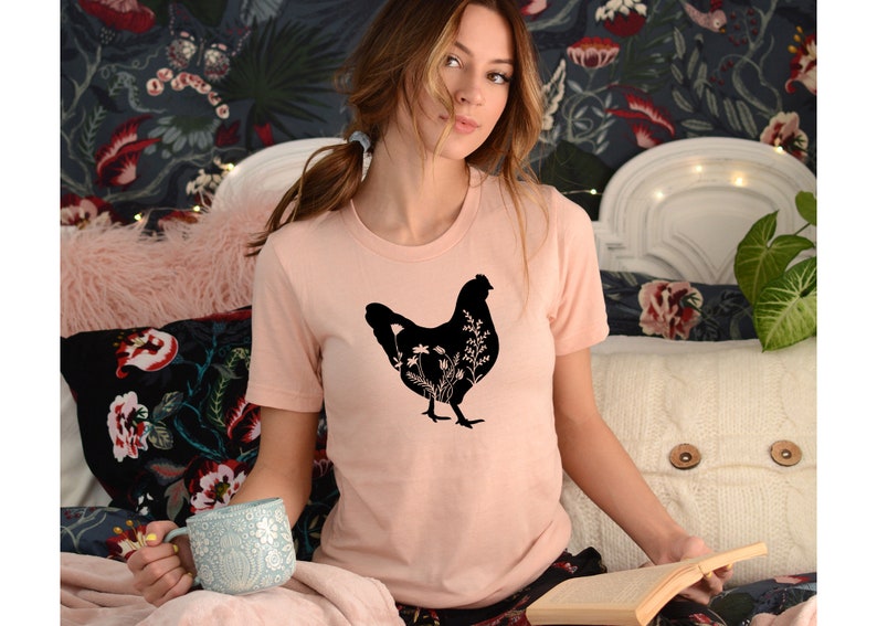 Pode incluir: Uma camiseta rosa com uma silhueta preta de uma galinha com flores dentro. A galinha est&aacute; virada para a direita e tem um pesco&ccedil;o comprido e penas de cauda.