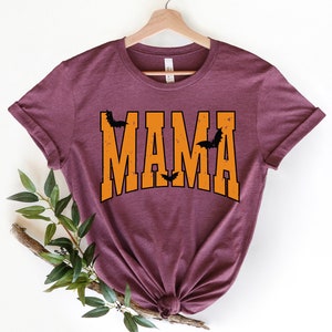 May include: A maroon t-shirt with the word "MAMA" in orange block letters. Black bat silhouettes are placed above and between the letters. The shirt is displayed on a wooden hanger.