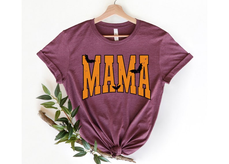 May include: A maroon t-shirt with the word "MAMA" in orange block letters. Black bat silhouettes are placed above and between the letters. The shirt is displayed on a wooden hanger.
