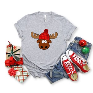 May include: A gray t-shirt with a cartoon moose wearing a red and white knit hat. The moose has brown antlers and a brown face with big eyes.