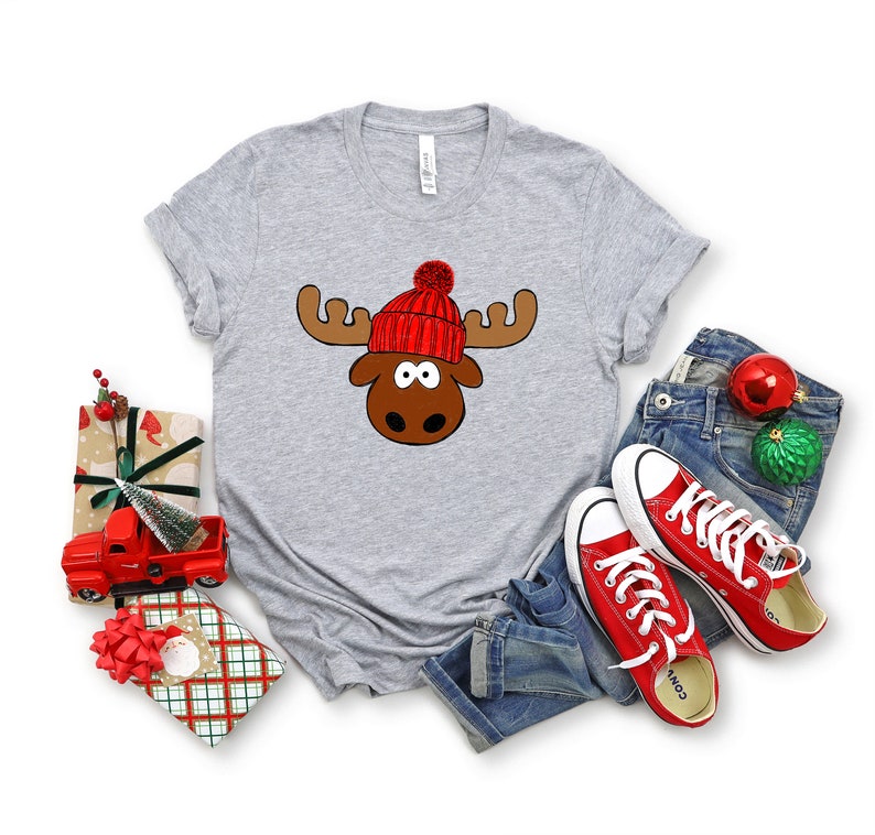 May include: A gray t-shirt with a cartoon moose wearing a red and white knit hat. The moose has brown antlers and a brown face with big eyes.