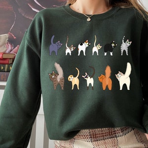 May include: A dark green sweatshirt with a print of multiple cats with their tails up in the air. The cats are in various colors and patterns.