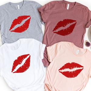 May include: Four t-shirts with a red lip print design. The shirts are in different colors: gray, maroon, white, and pink.