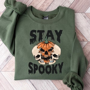 以下が含まれることがあります： オリーブグリーンのスウェットシャツ。黒い滴るフォントで「STAY SPOOKY」の文字。中央には、2つのドクロが付いたオレンジ色のカボチャ。ハロウィーンをテーマにしたスウェットシャツです。