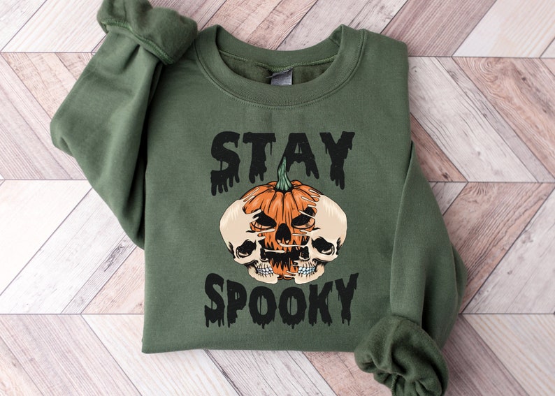 以下が含まれることがあります： オリーブグリーンのスウェットシャツ。黒い滴るフォントで「STAY SPOOKY」の文字。中央には、2つのドクロが付いたオレンジ色のカボチャ。ハロウィーンをテーマにしたスウェットシャツです。