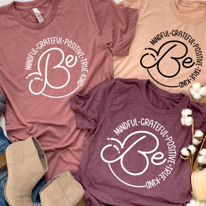 May include: Three t-shirts with the words "Mindful-Grateful-Positive-True-Kind" and a stylized "Be" in a circle. The shirts are in shades of pink and burgundy.