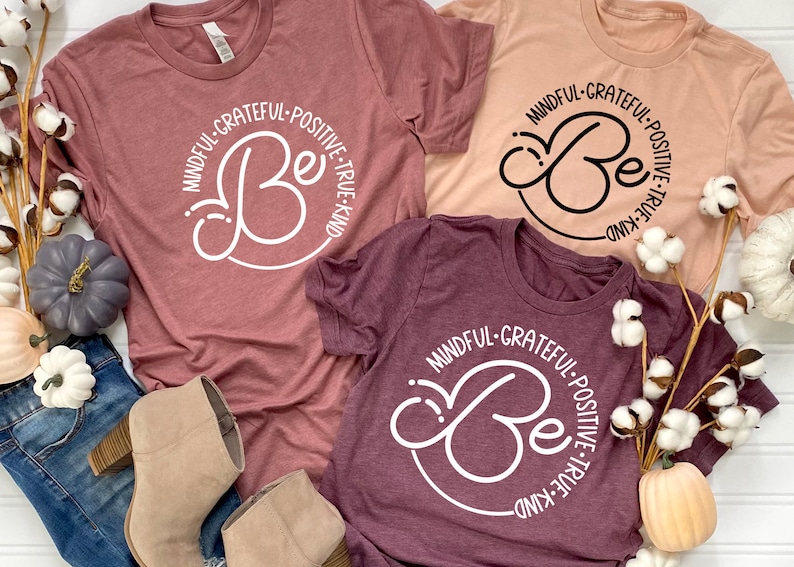 May include: Three t-shirts with the words "Mindful-Grateful-Positive-True-Kind" and a stylized "Be" in a circle. The shirts are in shades of pink and burgundy.