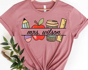 May include: A pink t-shirt with a graphic design of a red apple, a pencil, a coffee cup, and a ruler. The text "mrs. wilson" is printed on the shirt.
