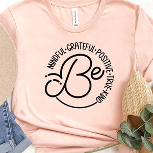 May include: A pink t-shirt with a black circle design that says "Be" in a cursive font. The circle is surrounded by the words "Mindful, Grateful, Positive, True, Kind".