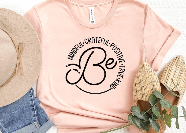 May include: A pink t-shirt with a black circle design that says "Be" in a cursive font. The circle is surrounded by the words "Mindful, Grateful, Positive, True, Kind".