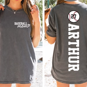 Puede incluir: Una camiseta gris con el texto "Baseball Mom" y un corazón en la parte delantera. La parte posterior de la camiseta tiene una pelota de béisbol blanca con el número 27 y el nombre "ARTHUR" impreso verticalmente en la parte posterior.