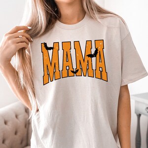 May include: Beige t-shirt with the word "MAMA" in orange block letters, with black bat silhouettes. The shirt has a distressed, vintage look, perfect for Halloween or a spooky theme.