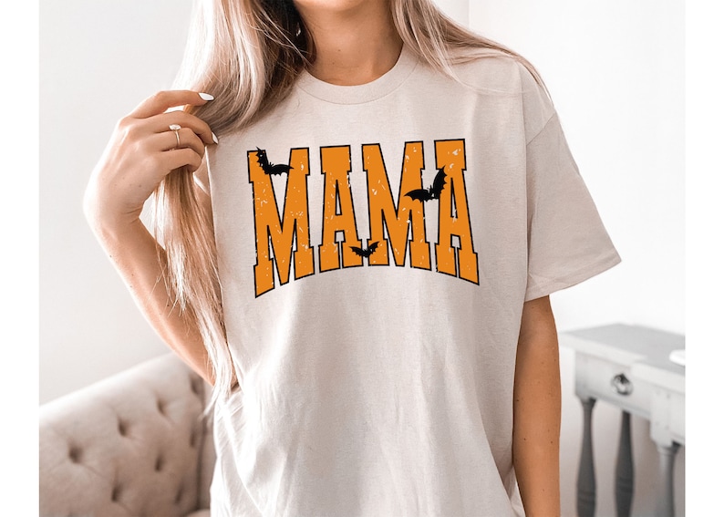 May include: Beige t-shirt with the word "MAMA" in orange block letters, with black bat silhouettes. The shirt has a distressed, vintage look, perfect for Halloween or a spooky theme.