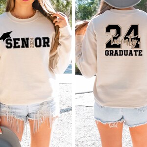 Puede incluir: Sudadera beige con texto negro y un gr&aacute;fico de birrete. La parte delantera dice "SENIOR 2024". La parte trasera presenta "24 Twenty GRADUATE" en letras negras y doradas. La sudadera es de estilo casual.
