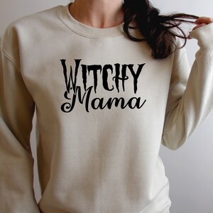 May include: A beige sweatshirt with the words "Witchy Mama" in black script. The text is centered on the front of the long-sleeved crew neck sweatshirt. The sweatshirt is a neutral color.