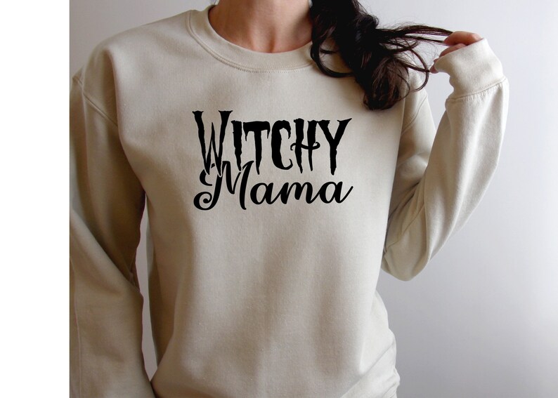 May include: A beige sweatshirt with the words "Witchy Mama" in black script. The text is centered on the front of the long-sleeved crew neck sweatshirt. The sweatshirt is a neutral color.